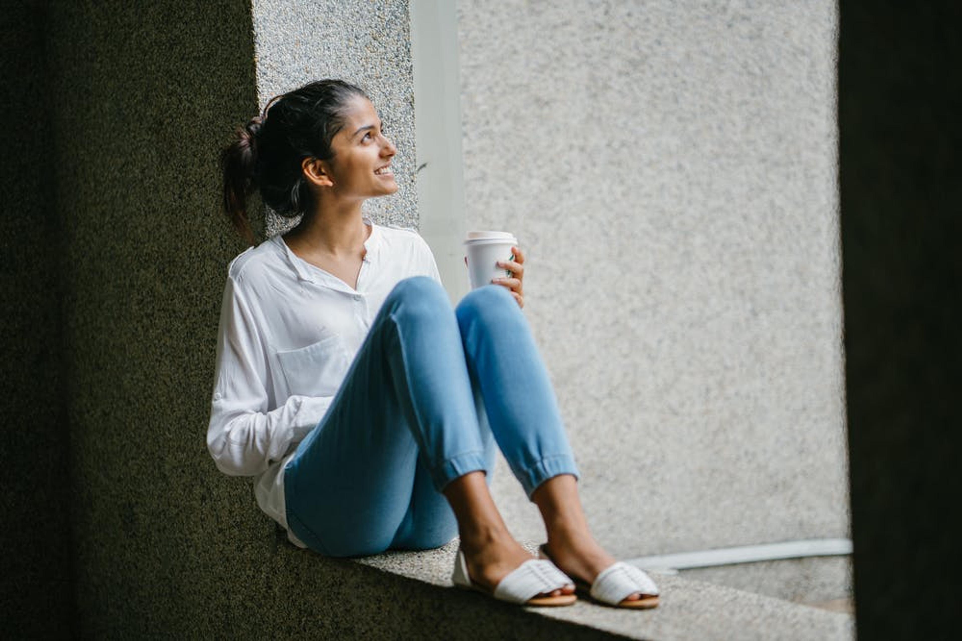 A young woman sits on a window ledge holding a cup of coffee, looking up and smiling