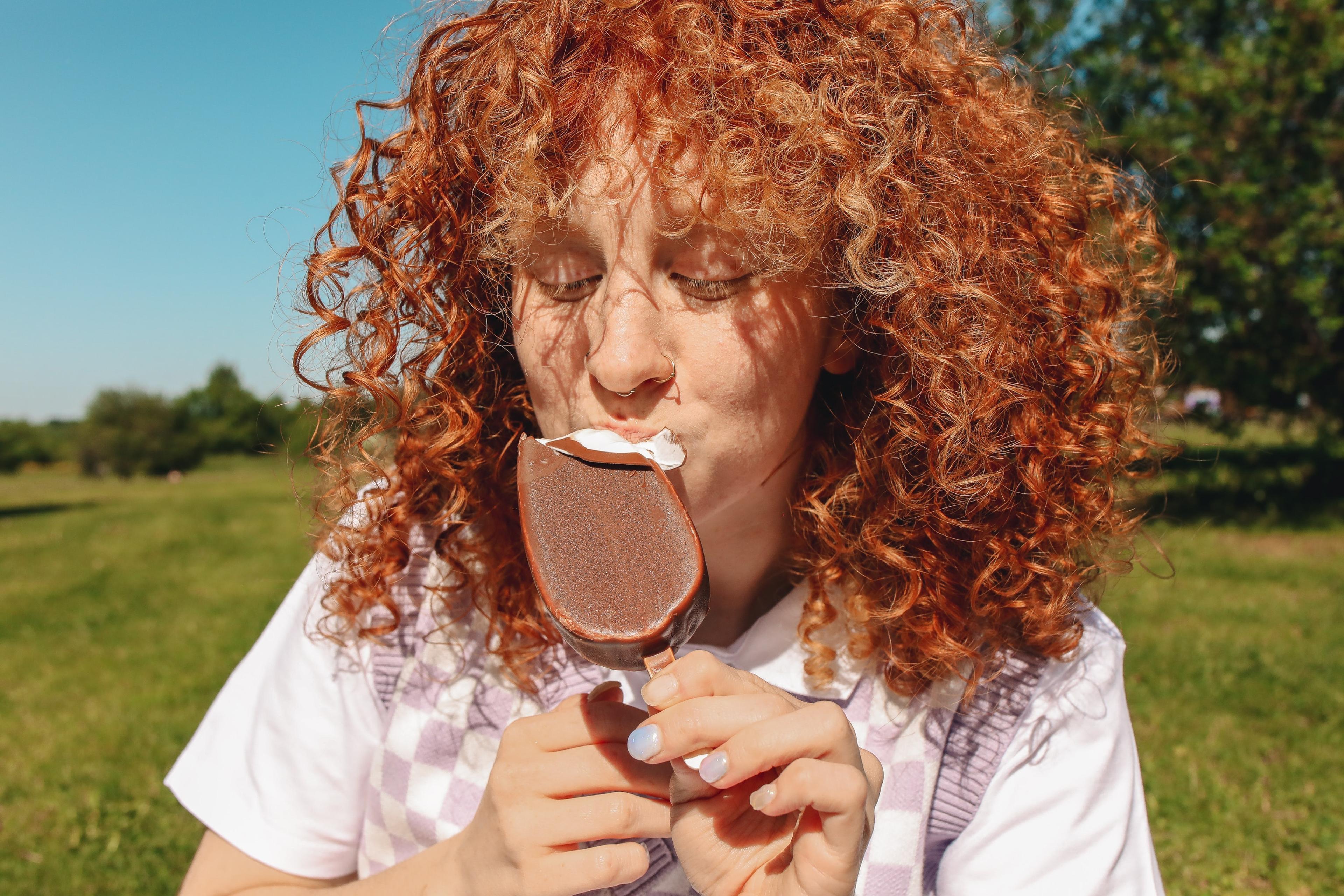 A woman with red curly hair eats an ice cream treat outside in the sun