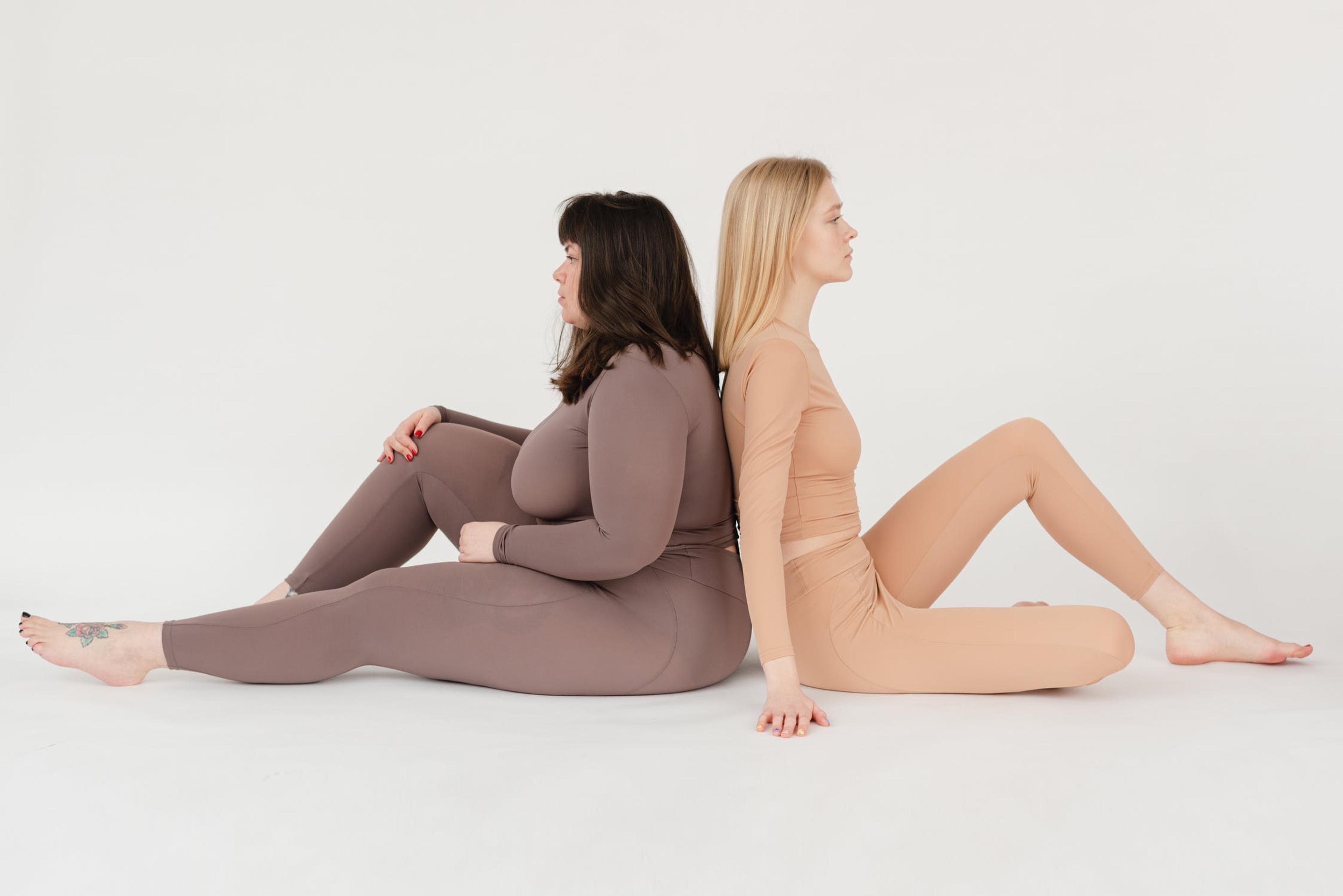 Two women with noticeably different body shapes and sizes sit back-to-back