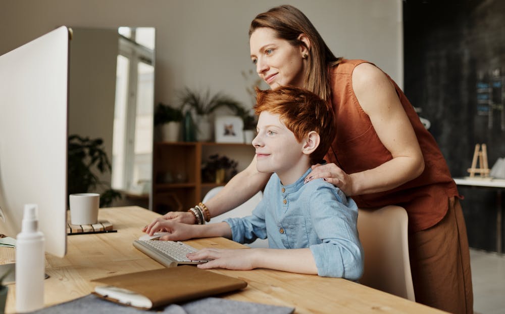 A smiling mom looks over her young son's shoulder while he works at the computer