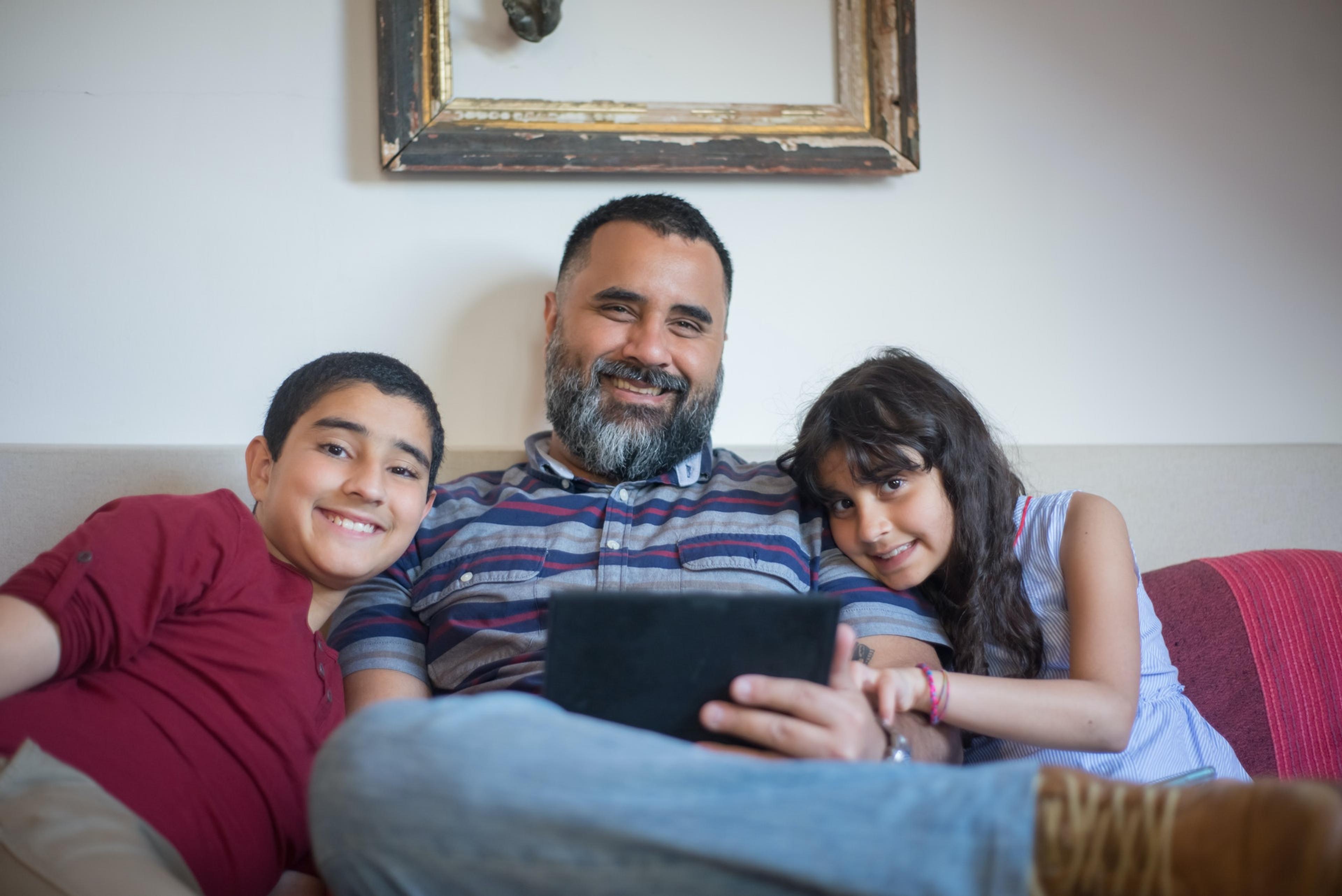 A smiling father sits on the couch between his young son and daughter, also smiling