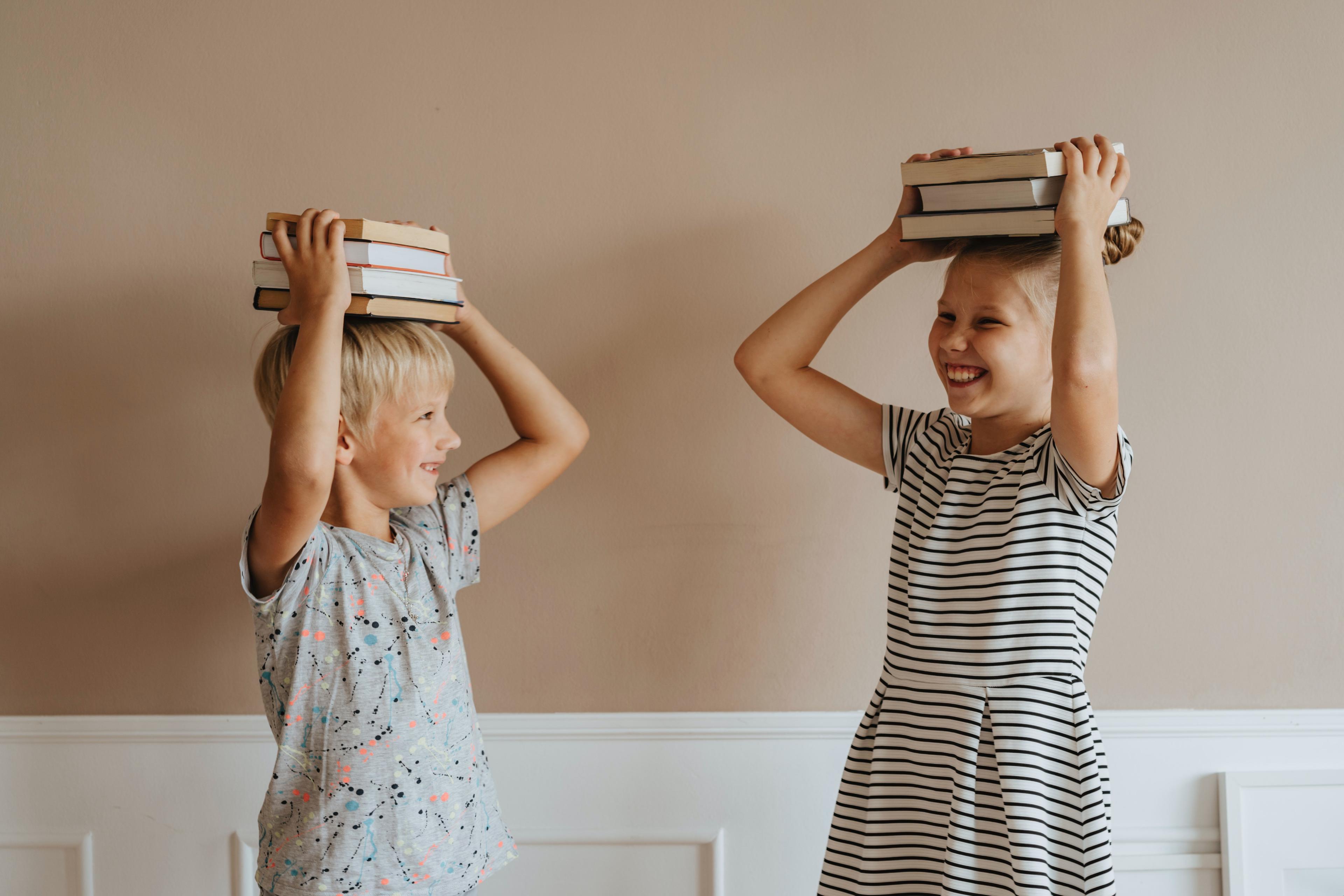 Two blond kids in t-shirts, holding books on top of their heads and laughing