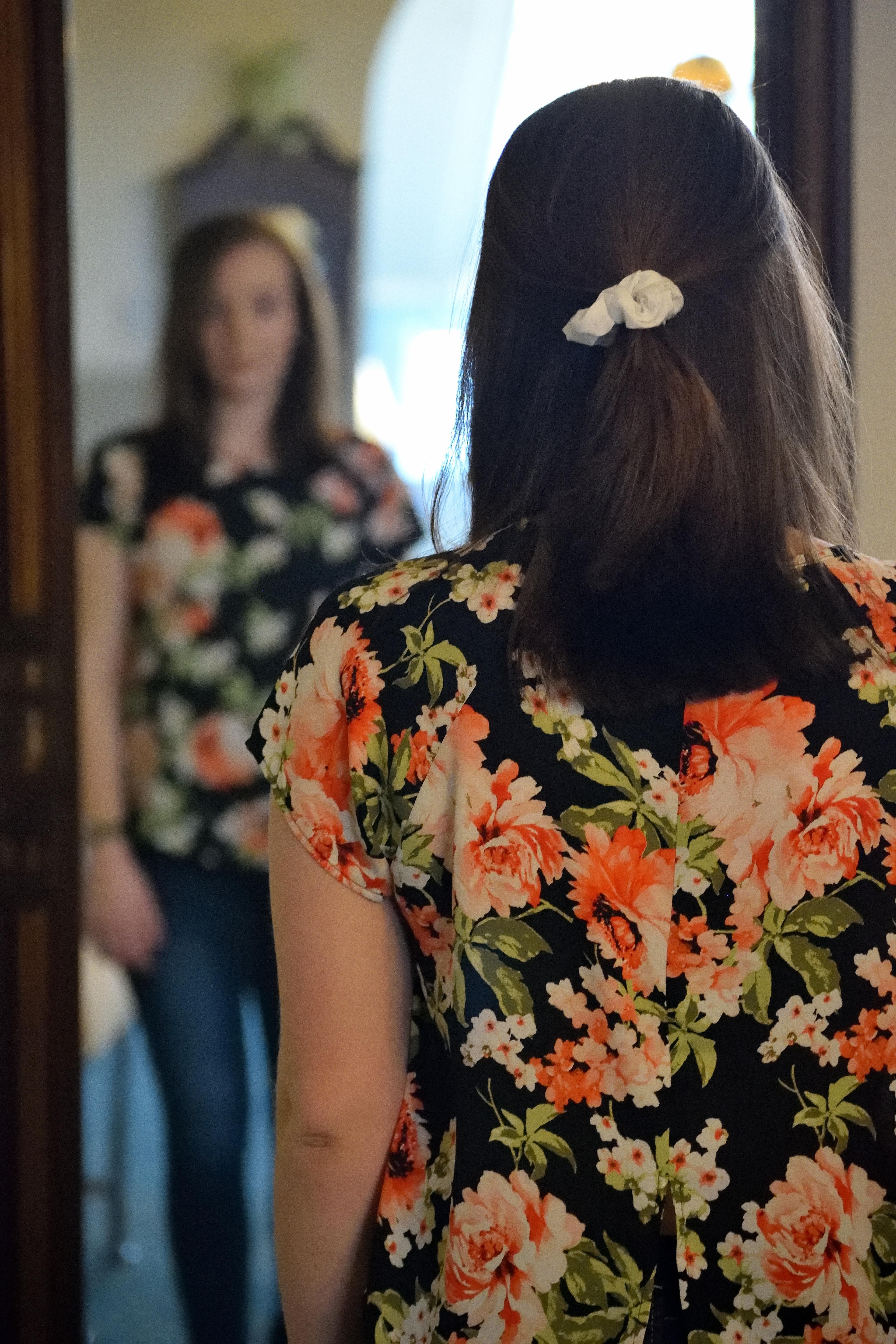 A young woman in a flowered shirt looks at her reflection in the mirror
