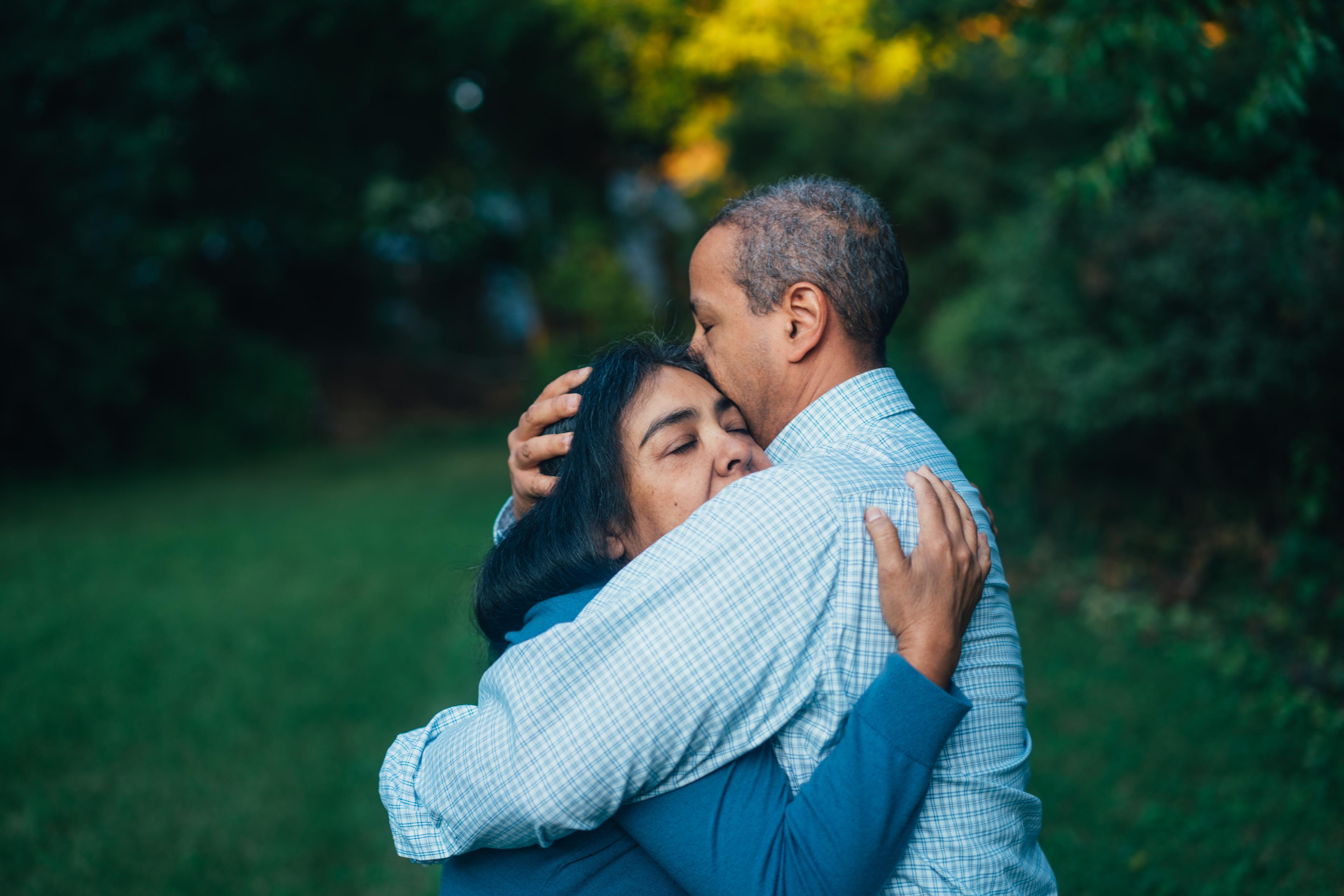 A man and a woman hug one another while outside in a green yard