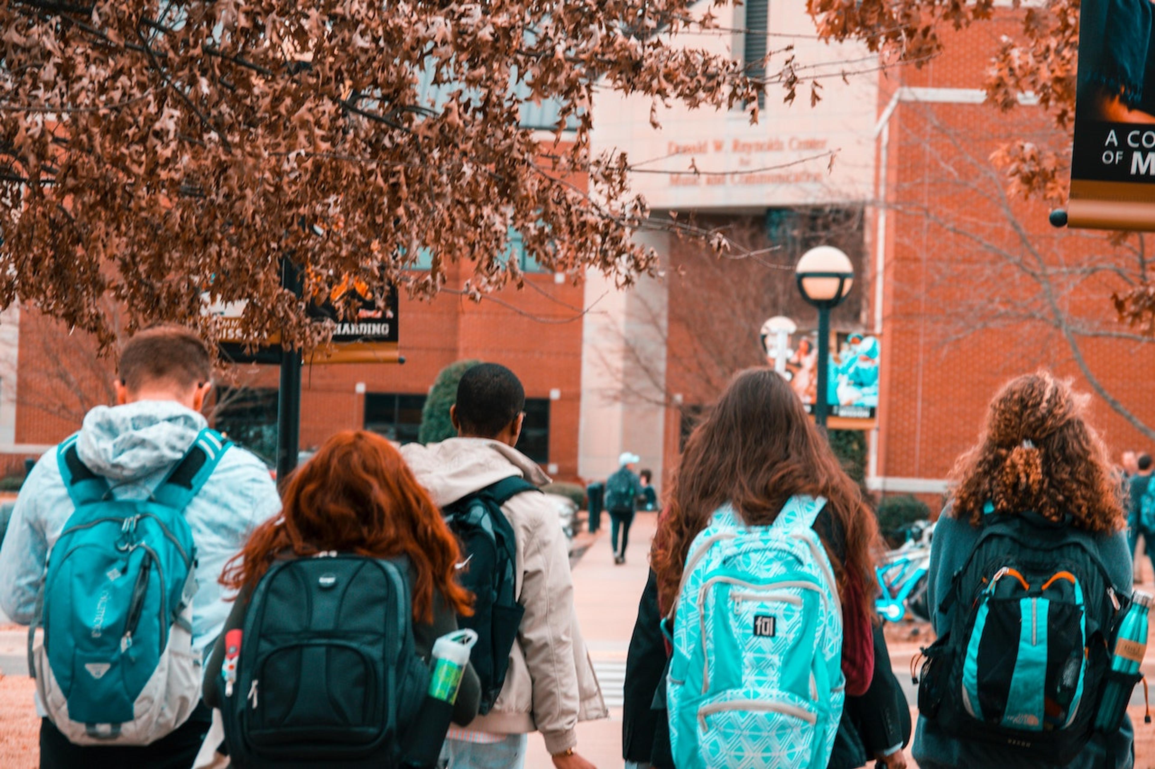 Several students, all wearing backpacks, walk toward a red brick building on a fall day