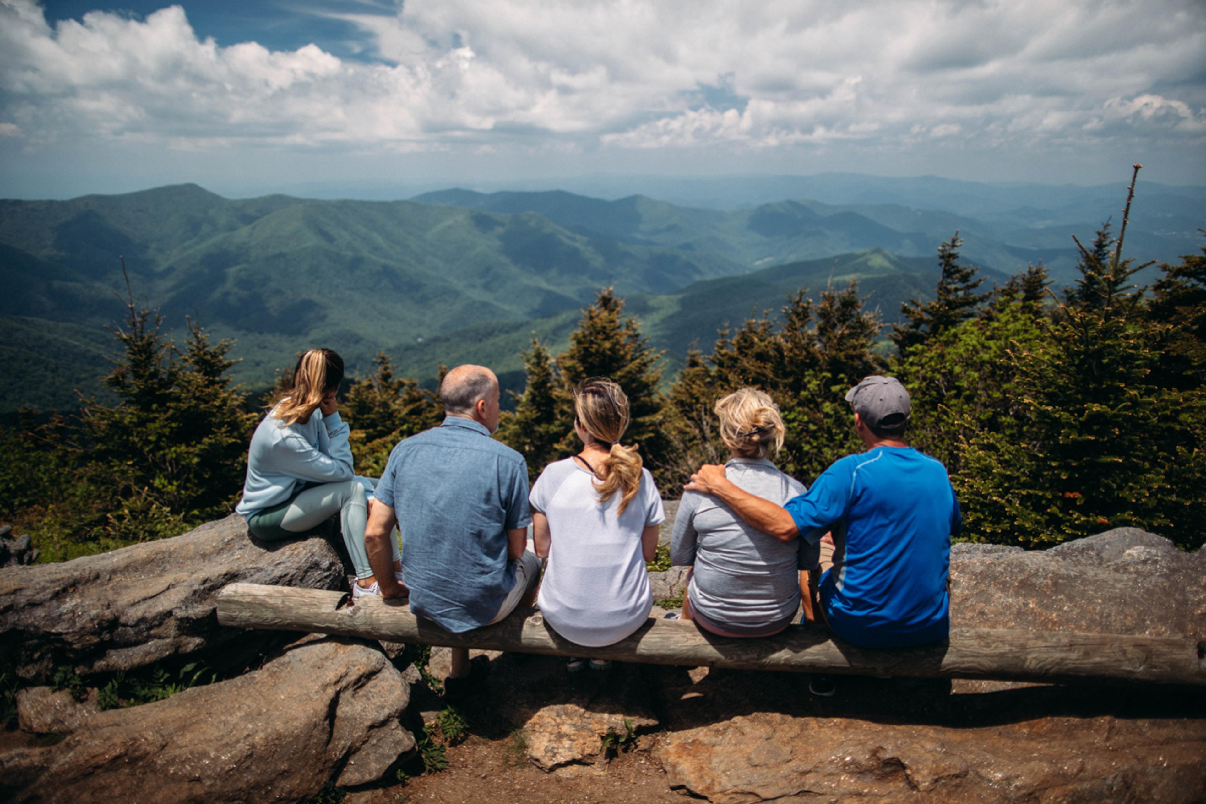 A family siting on a mountaintop, backs to the camera, looking out on a mountain range.