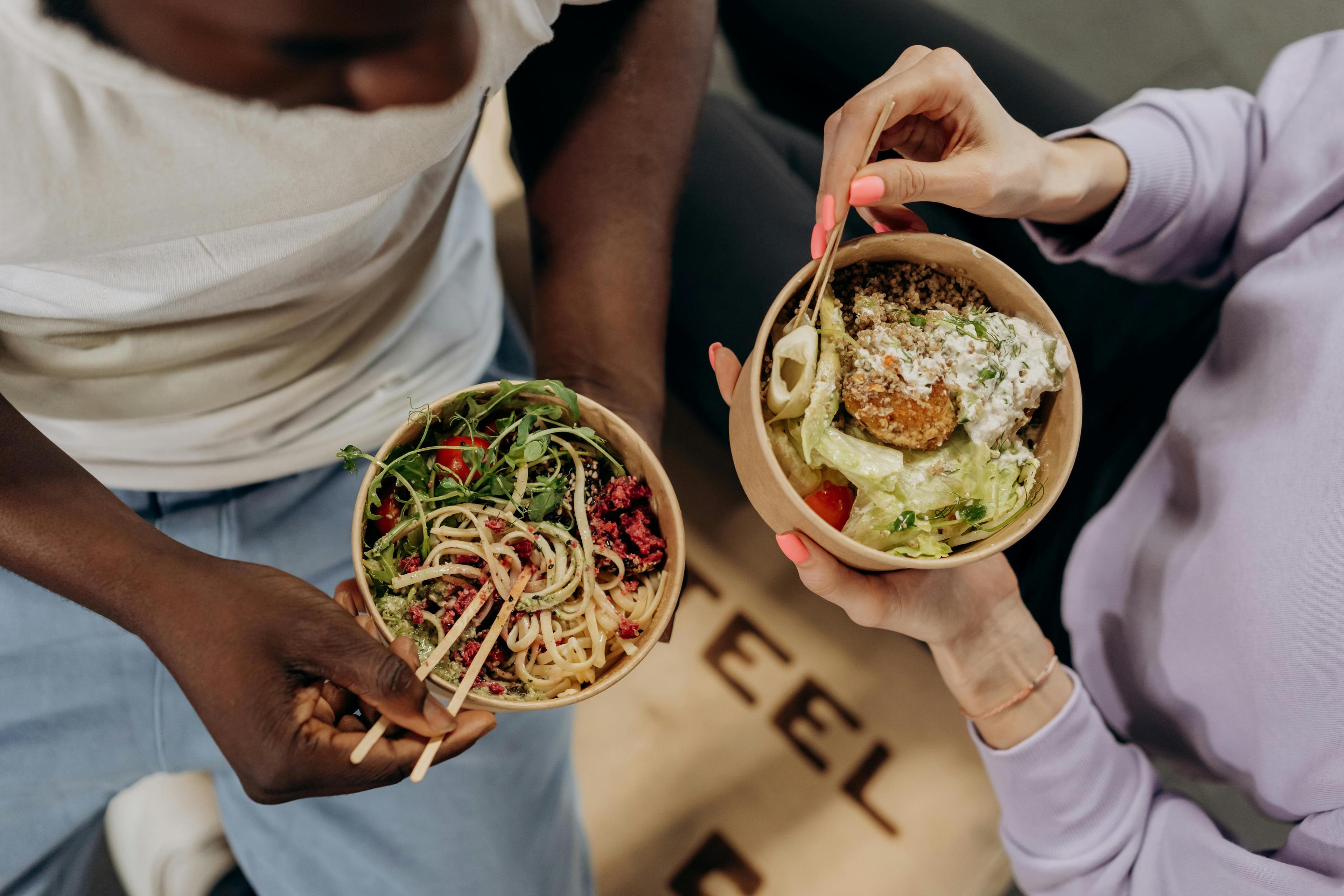 Overhead shot of two people eating bowls of different food in takeout containers