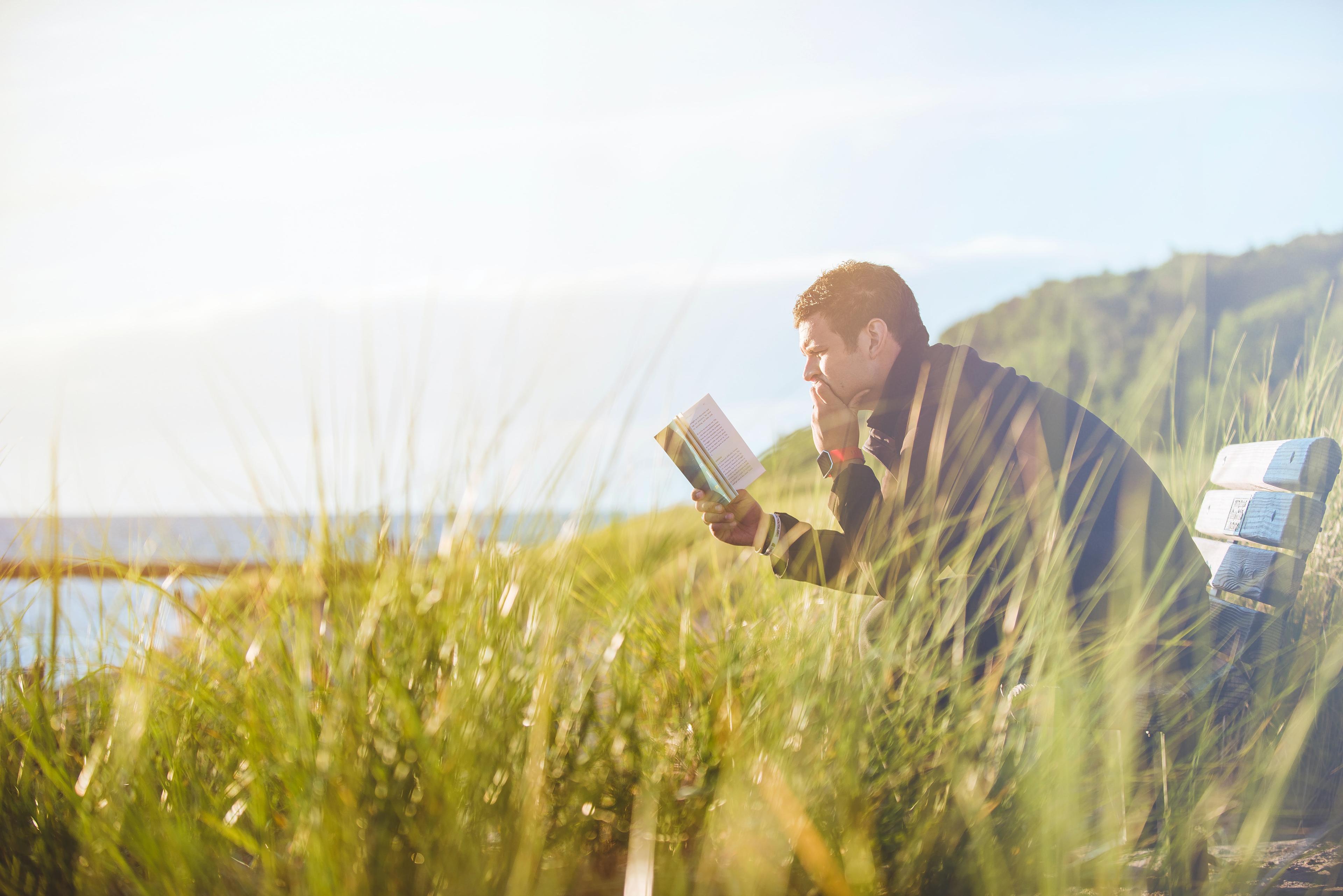 A man sits on a bench in tall grass by water, reading a book