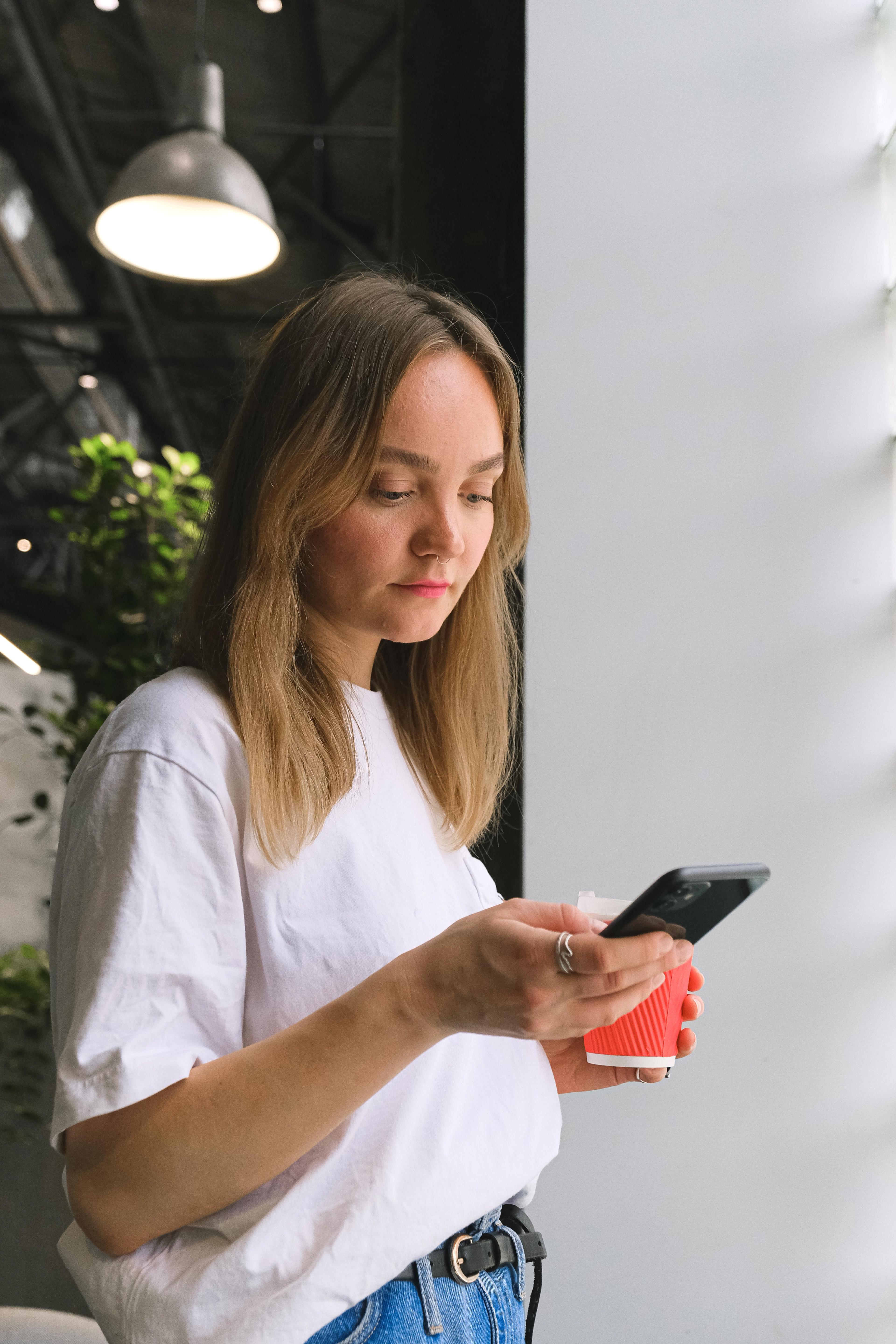 A woman in a white t-shirt and jeans holds a cup of coffee and looks at her phone