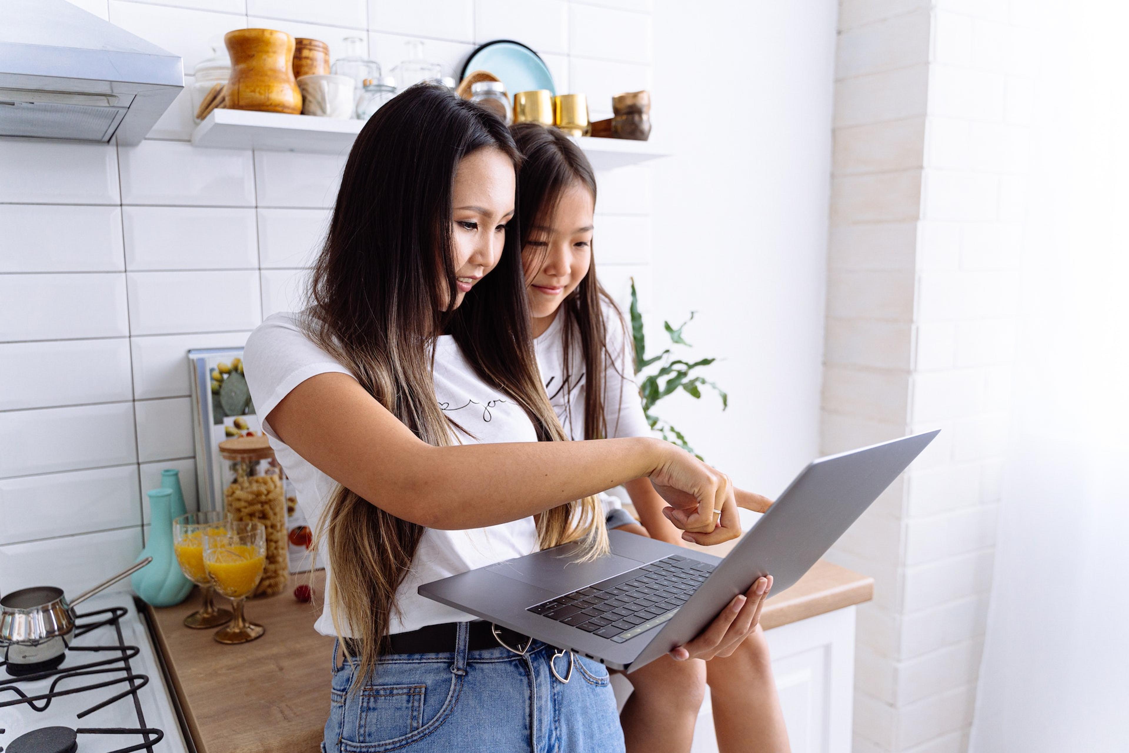 A young woman and a girl stand together in a kitchen looking at a laptop