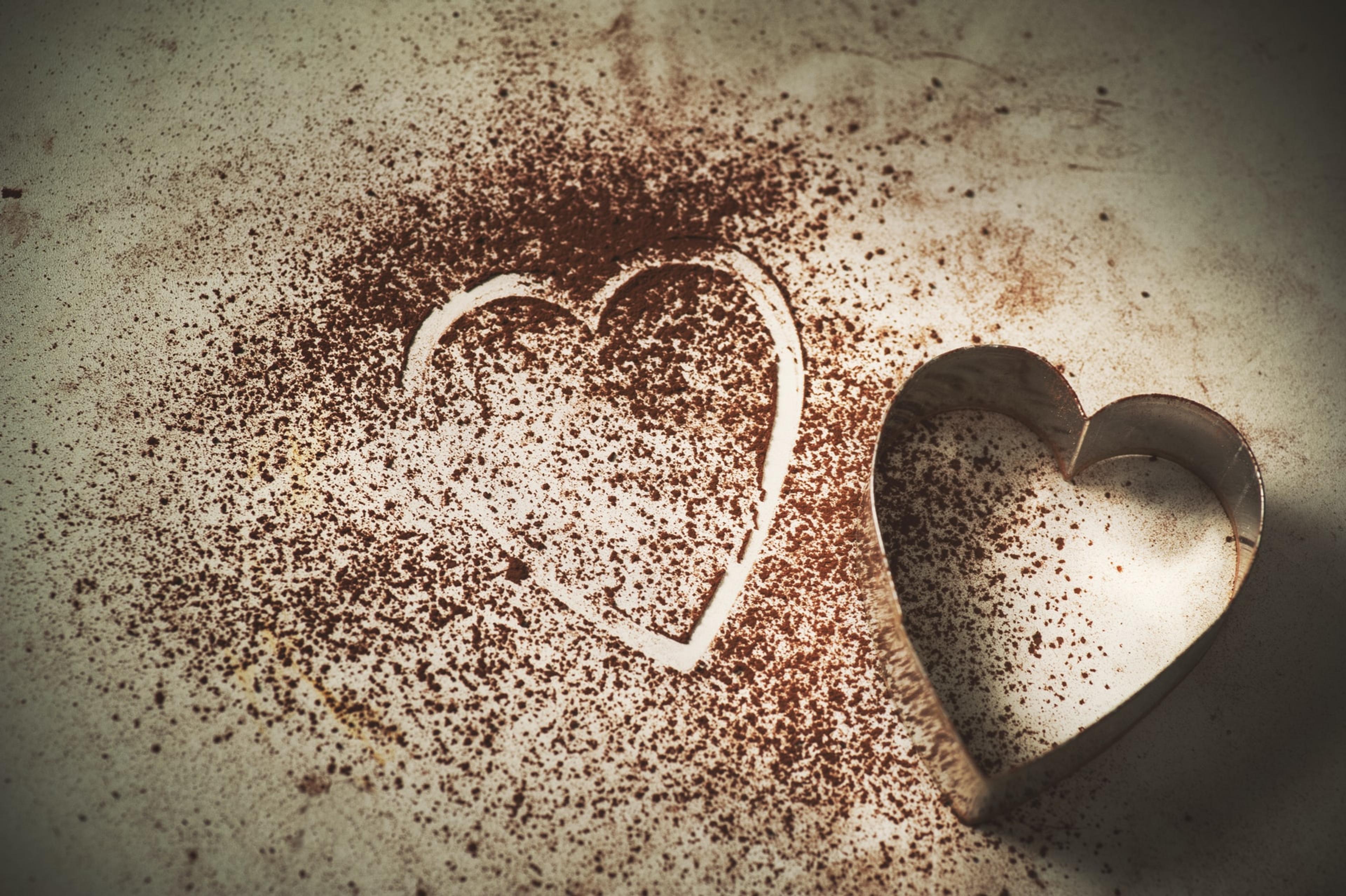 A heart-shaped cookie cutter on a surface next to spilled cocoa powder with a heart-shaped outline in it