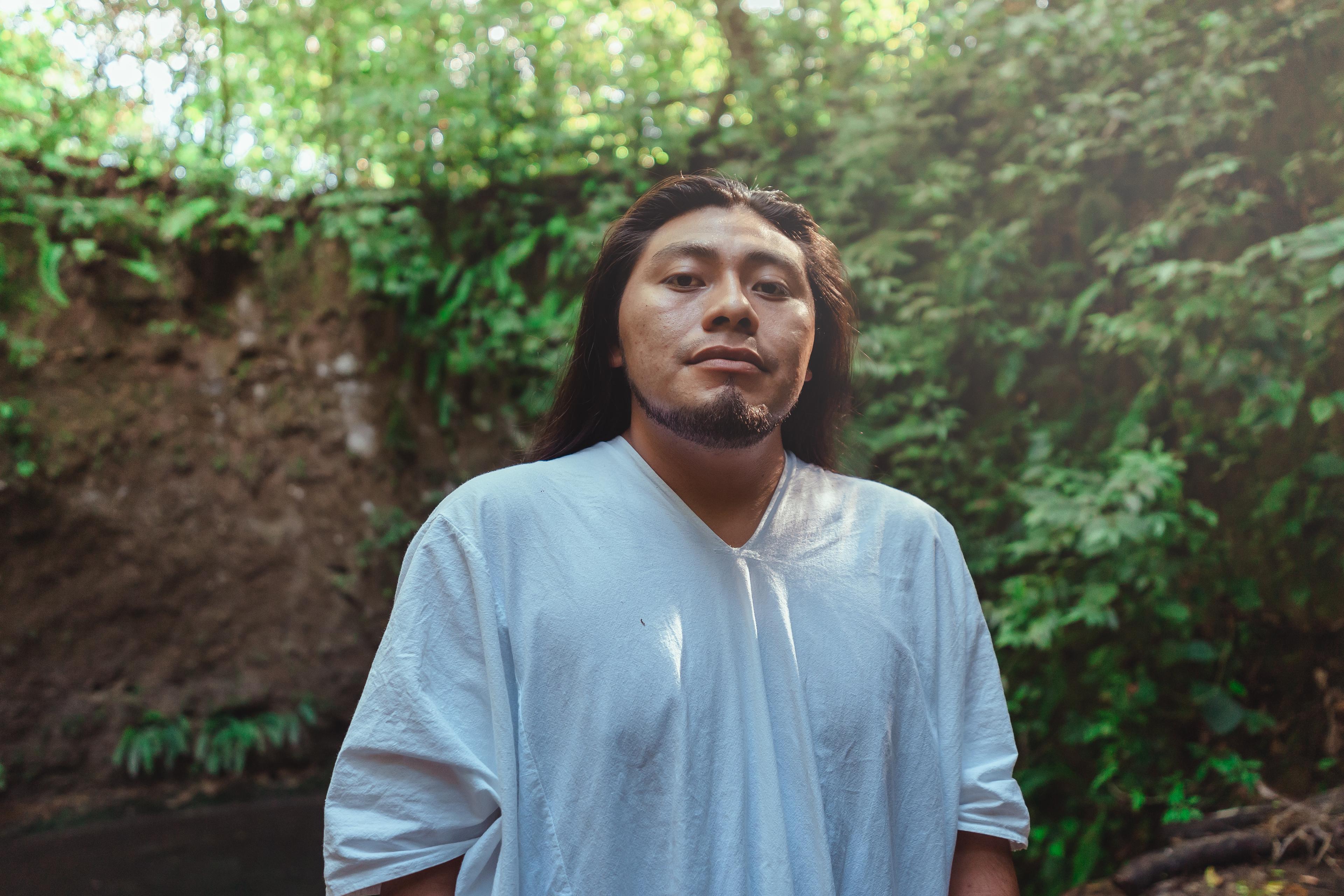 A Latinx man with long hair stands outside in nature