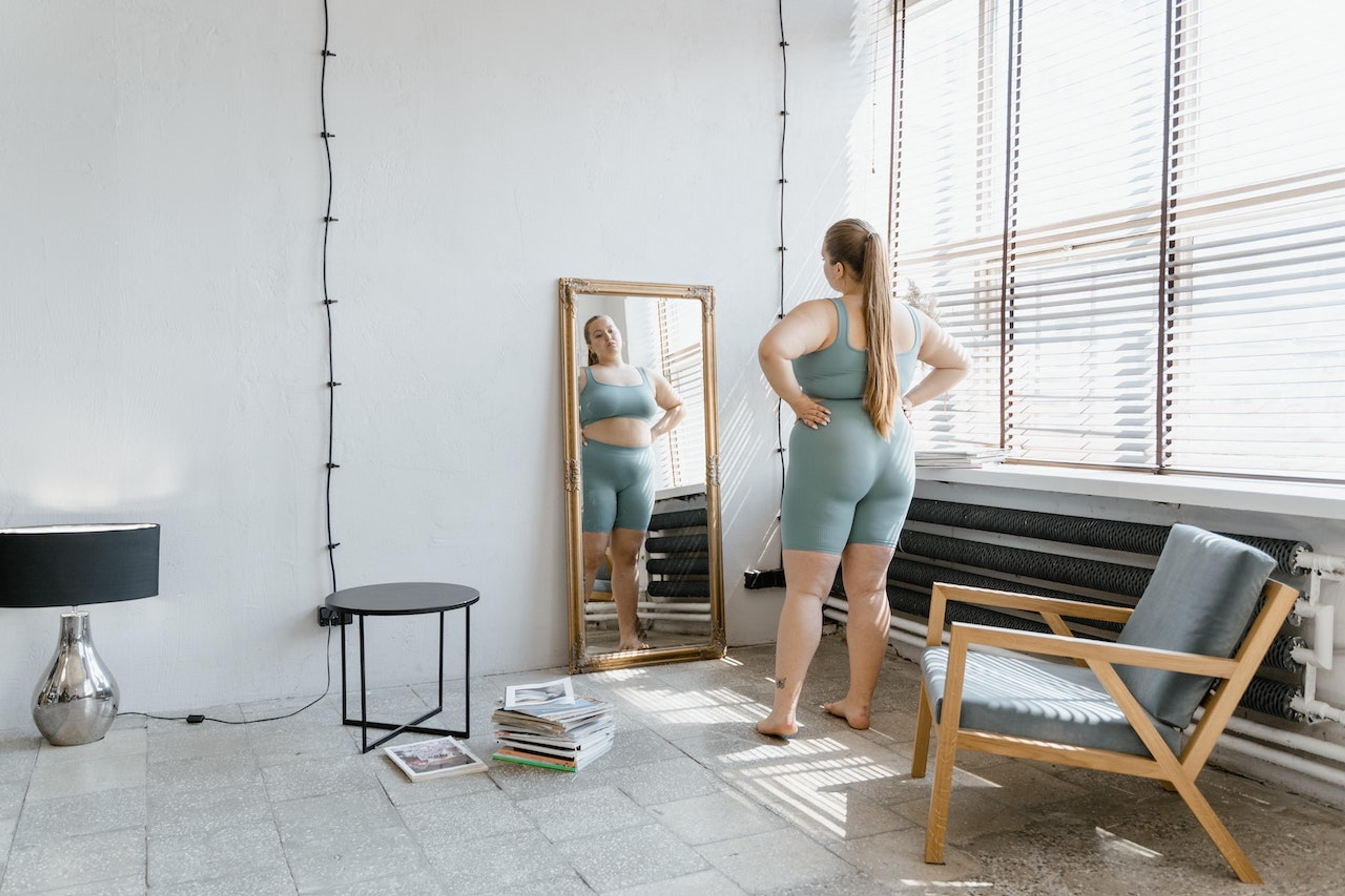 A woman in a larger body wearing matching sports bra and bike shorts looks at herself in the mirror