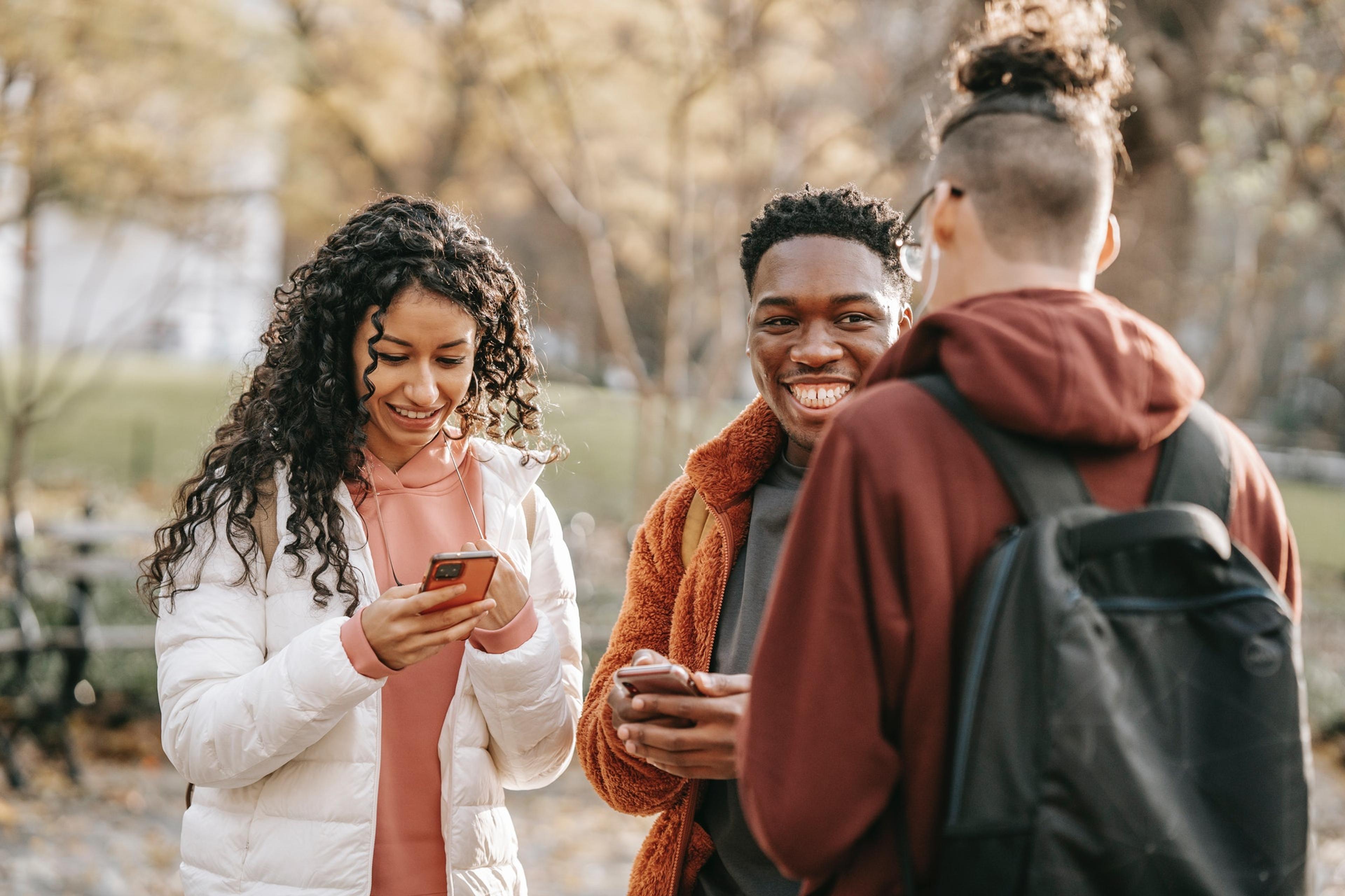 A group of three young adults stand together on a college campus holding their phones and smiling