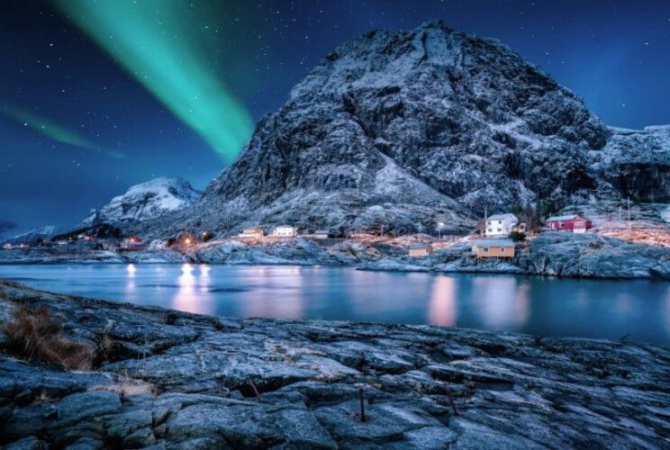 A snowy mountain with the northern lights behind a beautiful flowing sea next to Vesteralen in Norway.