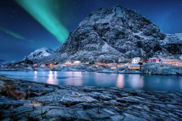 A snowy mountain with the northern lights behind a beautiful flowing sea next to Vesteralen in Norway.