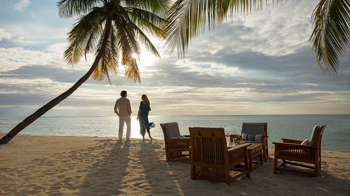 A couple stands on a peaceful beach at Paradis with a glorious sunset in the background.