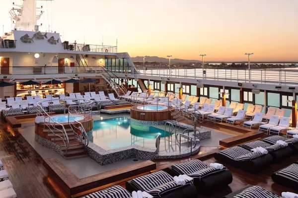 The pool deck aboard the Oceania Insignia with a clean pool with the beautiful sunset in the background, seats to relax in, a wonderful view of the ocean and a view of an island