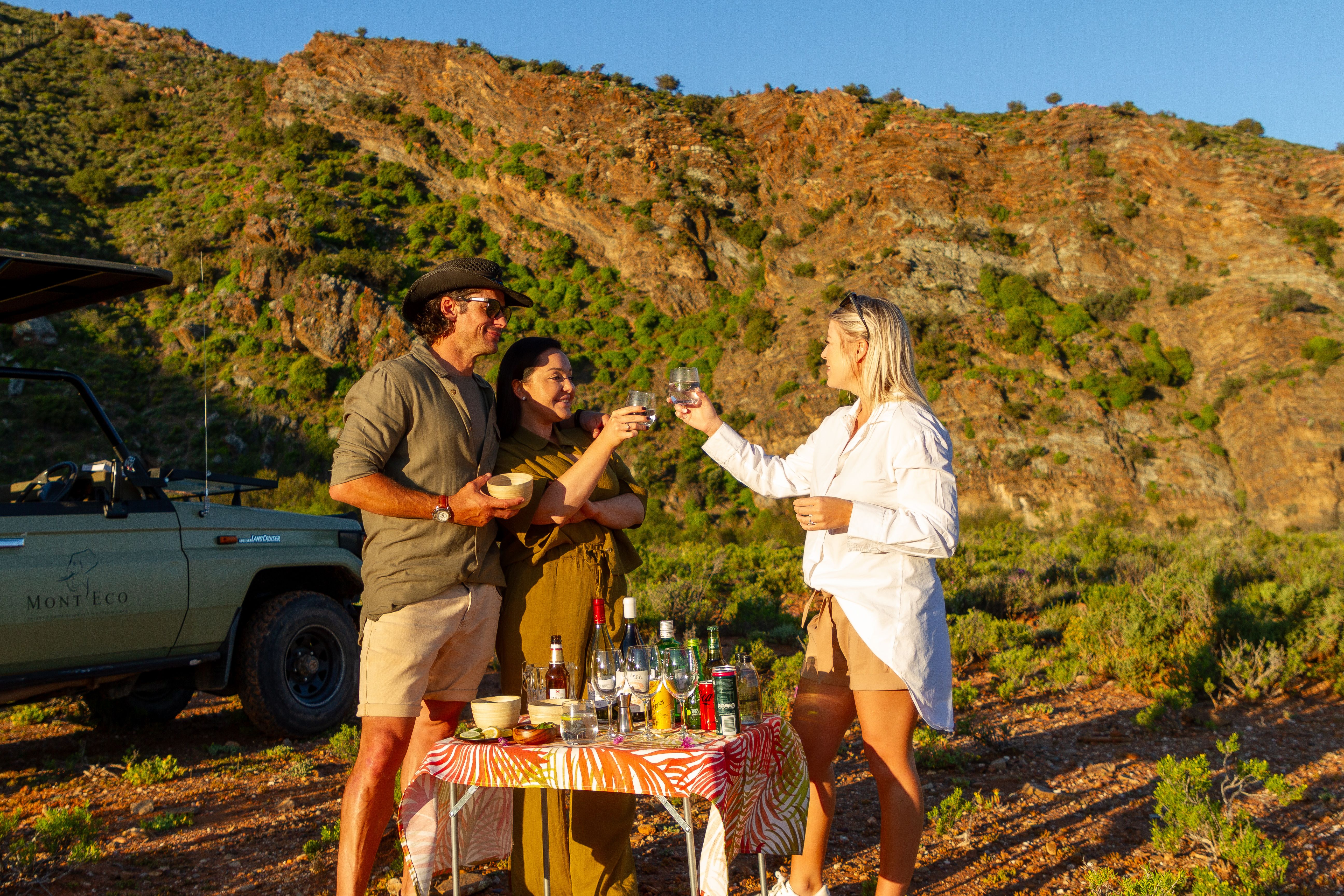 3 happy people raise a toast while having sundowner drinks in the bush on safari