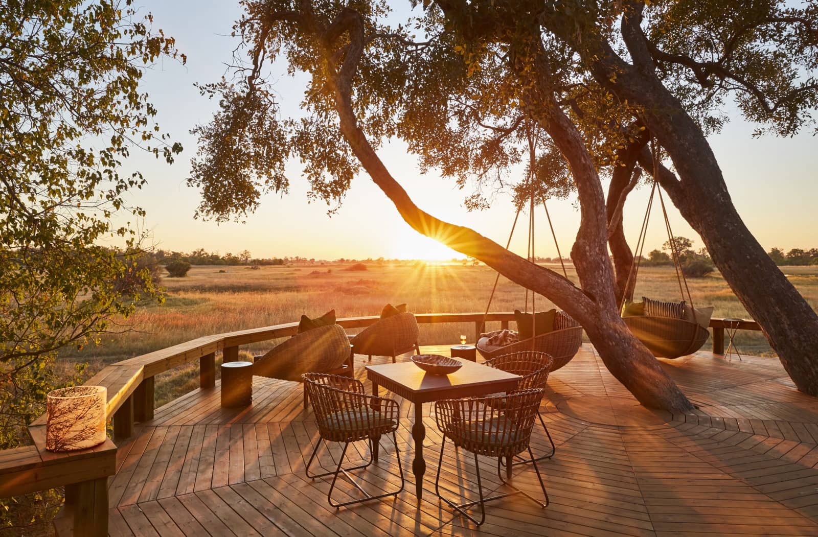 Wooden deck with table chairs loungers beneath a tree with sunsetting behind view of Okavango Delta