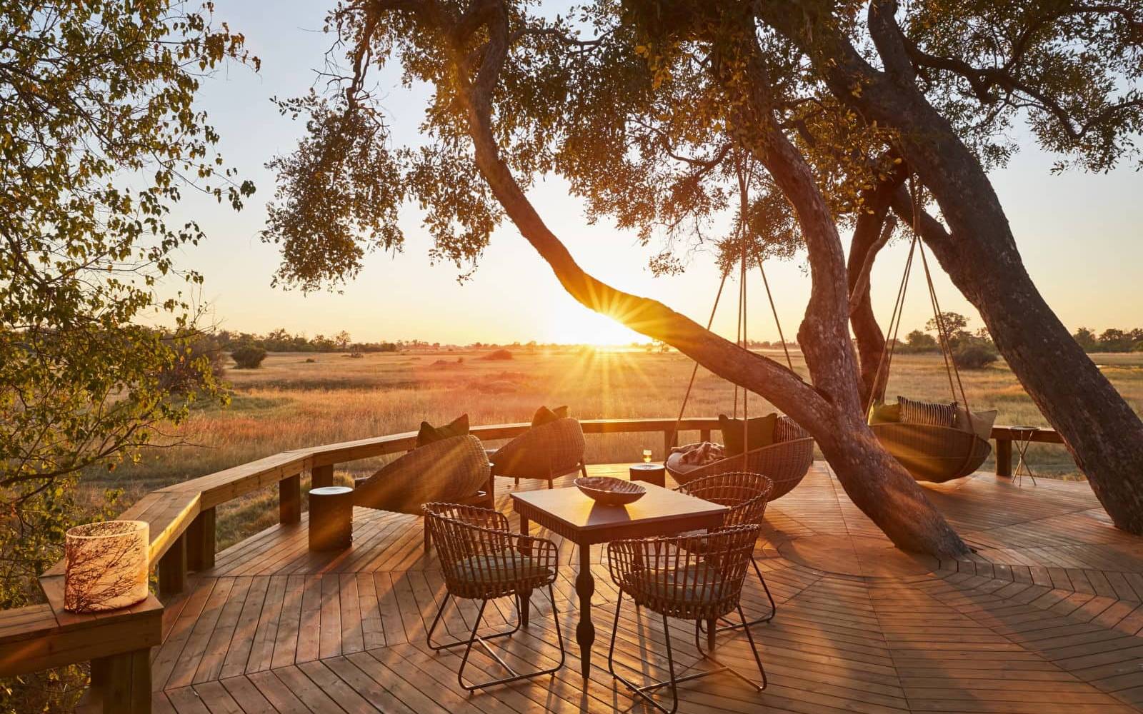Wooden deck with table chairs loungers beneath a tree with sunsetting behind view of Okavango Delta