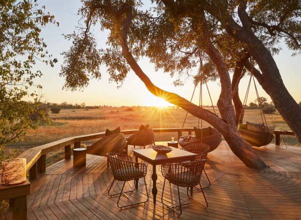 Wooden deck with table chairs loungers beneath a tree with sunsetting behind view of Okavango Delta