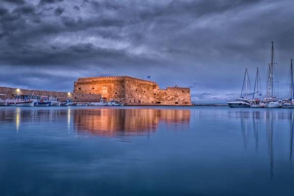 Majestic view of Koules Fortress reflected in calm waters at Heraklion harbor during twilight.