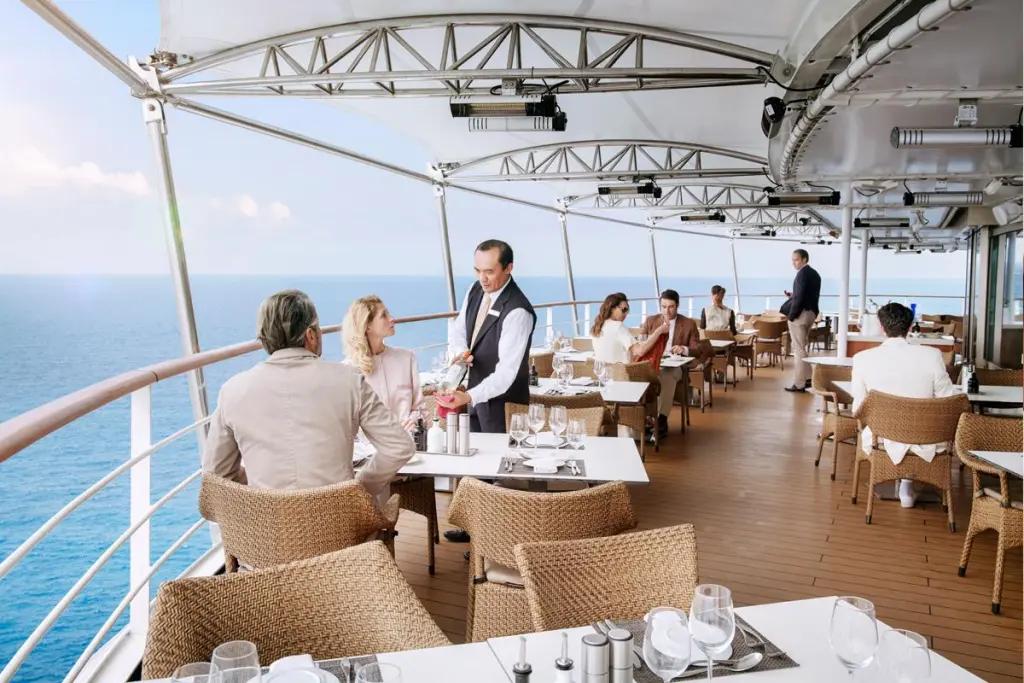 A couple sit at a table on the deck of the ship while they consider the wine presented by the smartly dressed sommelier.