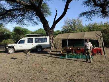 Male ranger standing in front of dining tent under a tree with game driving car parked beside