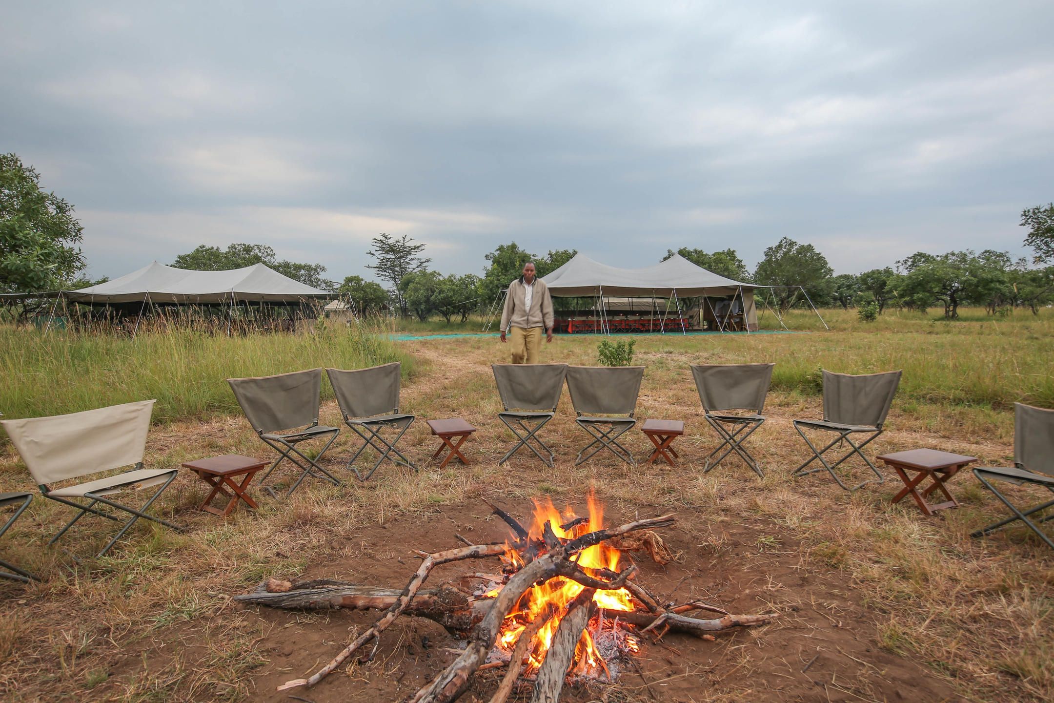 Campfire surrounded by camp chairs with person walking from two tents in the background