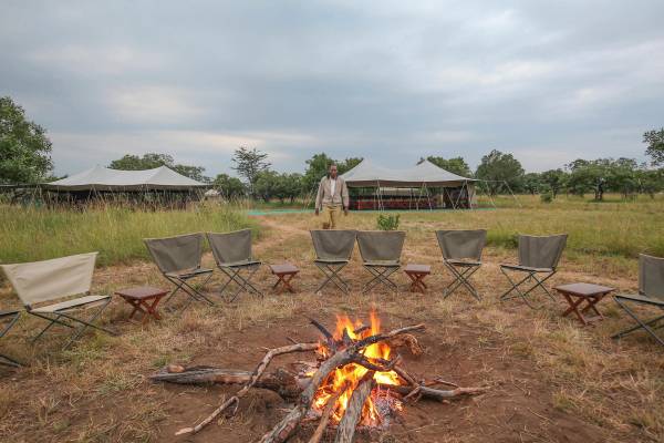 Campfire surrounded by camp chairs with person walking from two tents in the background