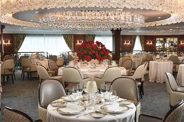 A picture of the Grand Dining Room aboard the Oceania Insignia with seats that look like silver, a table with lots of lovely flowers in the middle and wonderful looking cutlery.