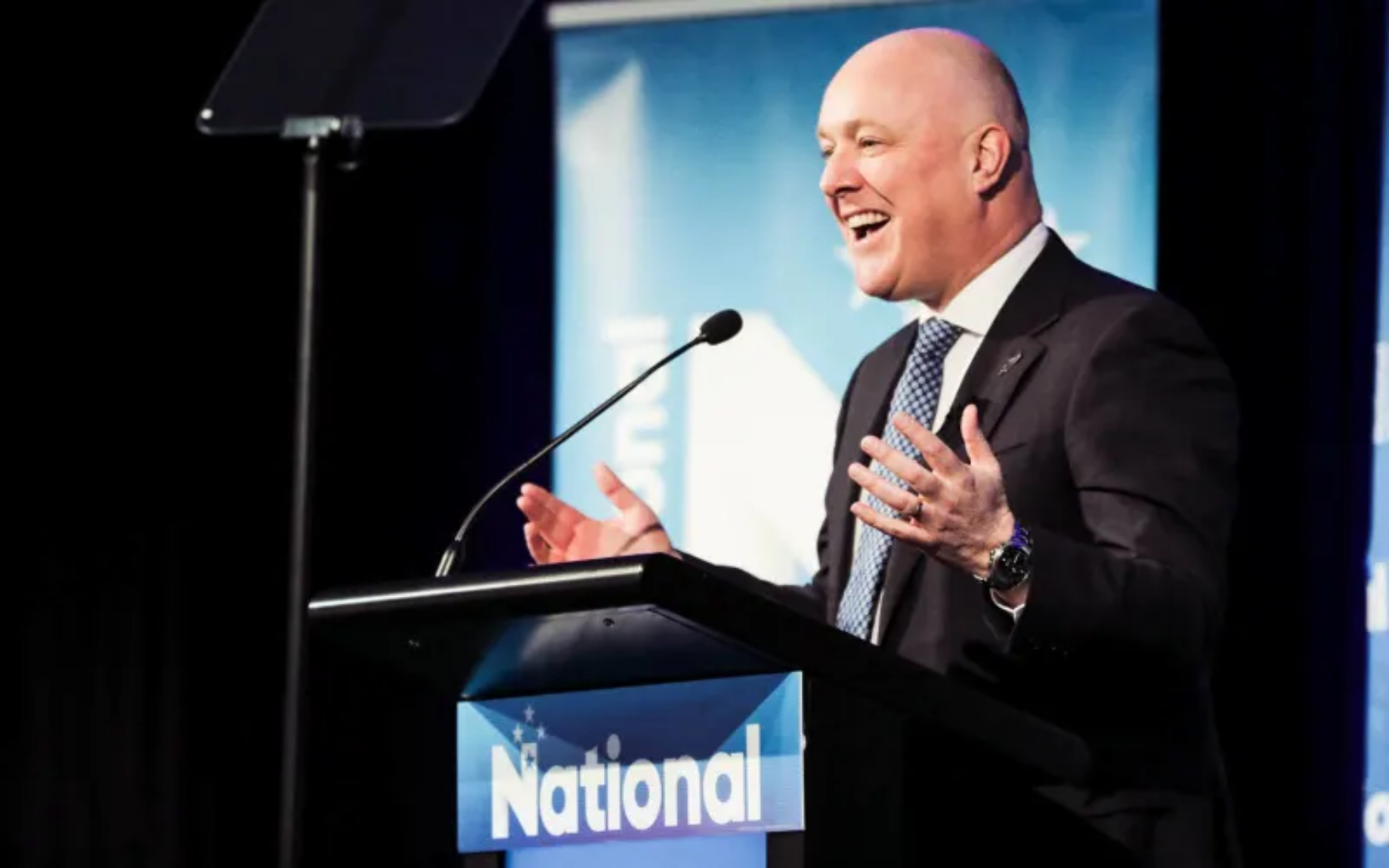 Christopher Luxon laughing and gesturing at a National Party podium