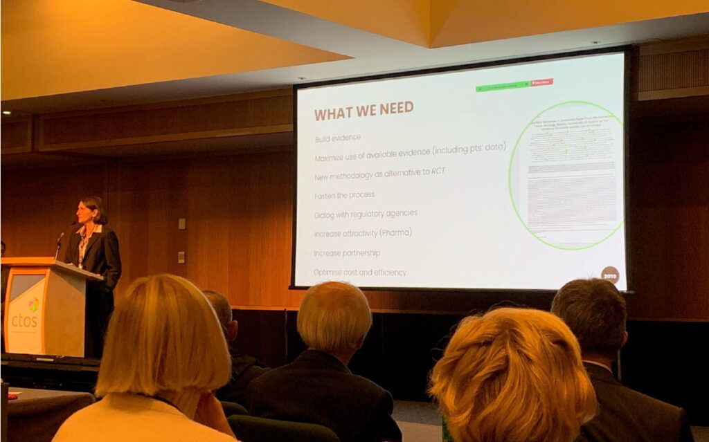 A presenter at the podium with a slide behind them titled: What we need