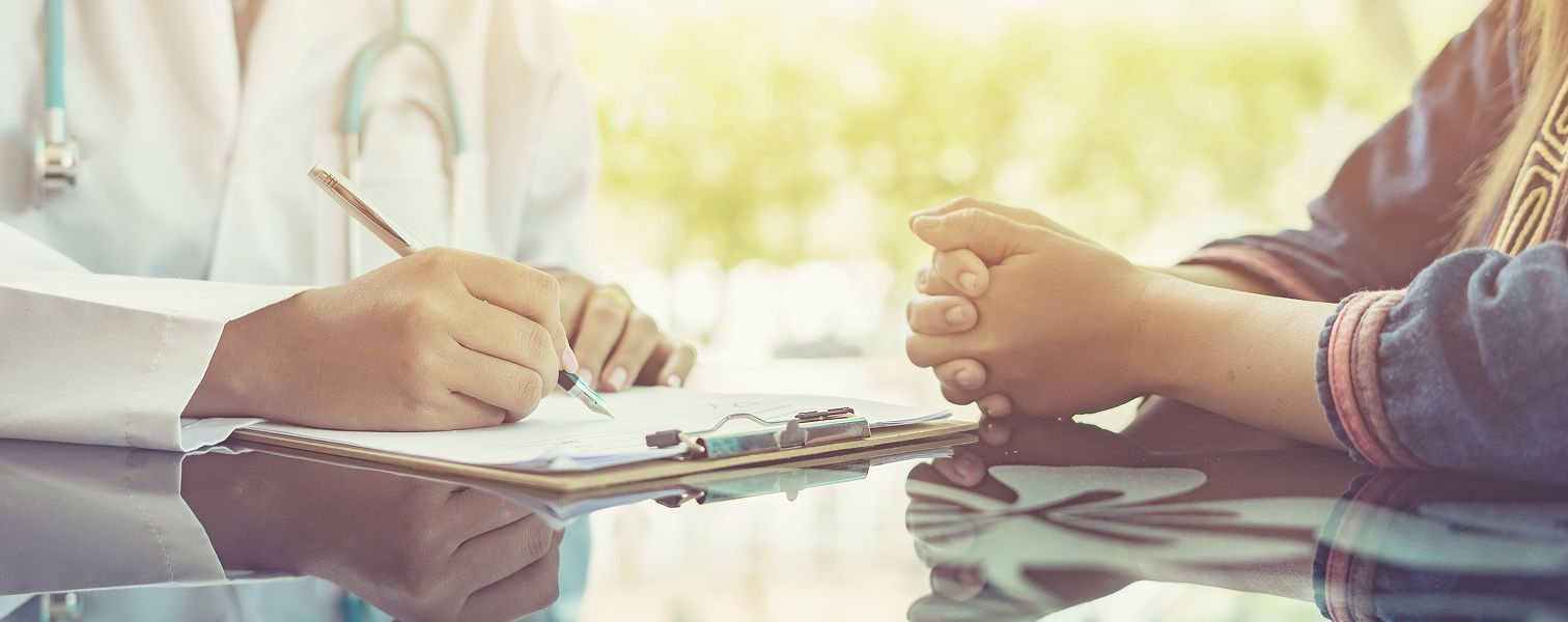 On the left, a sitting doctor is writing and on the right, a patient is also sitting with their hands together.