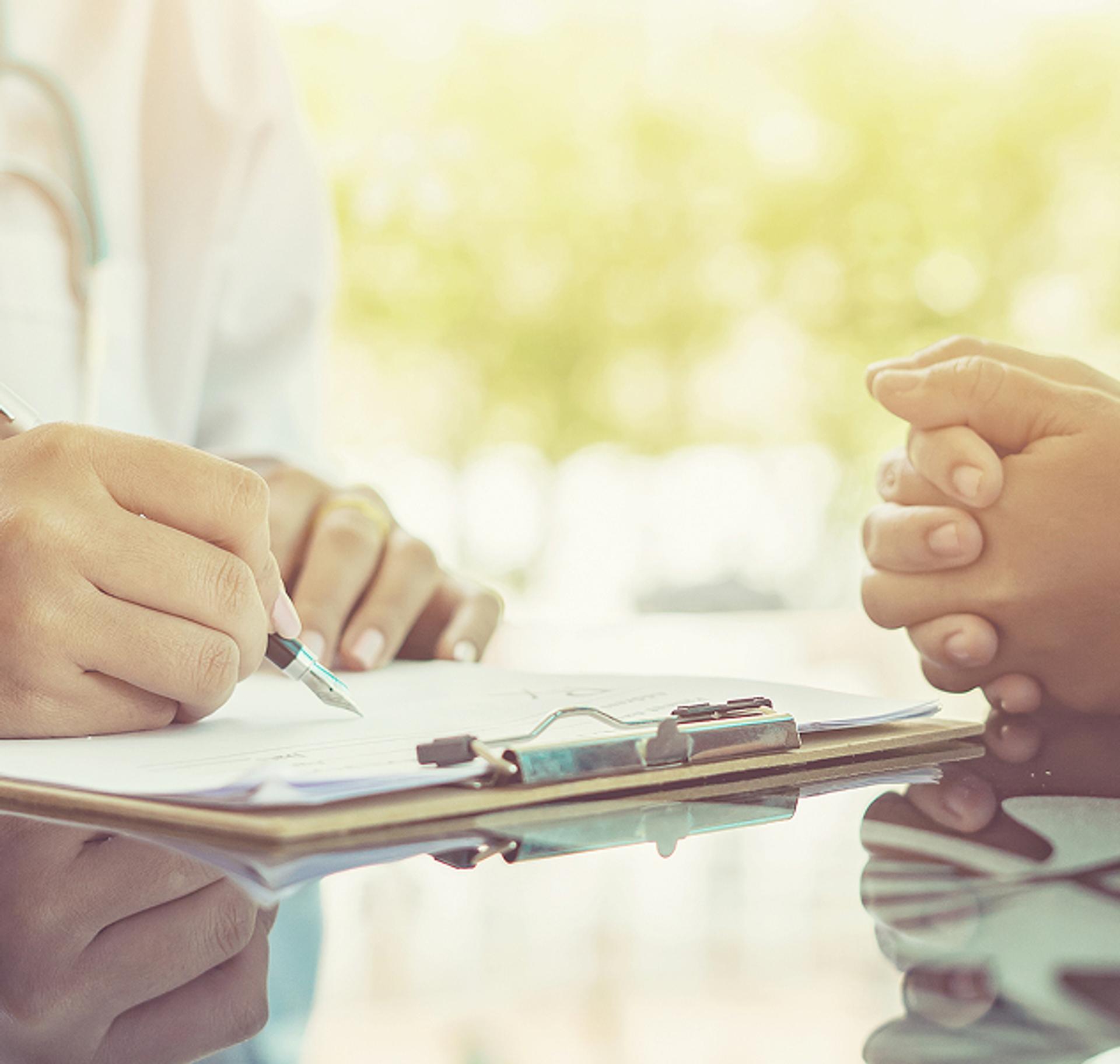 On the left, a sitting doctor is writing and on the right, a patient is also sitting with their hands together.
