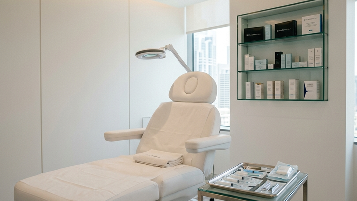 Luxury medical aesthetics treatment room with a white upholstered treatment chair, premium skincare products on a floating shelf, and subtle LED accent lighting, completely empty with no client or technician, communicating the booking gap