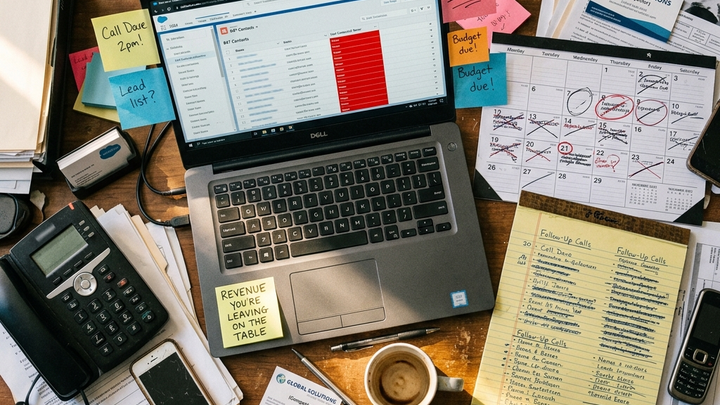 Top-down overhead photograph of a cluttered service business desk: phones, a laptop showing a CRM with 847 contacts and a Last Contacted Never column in red, handwritten follow-up lists, a scratched calendar, sticky notes, cold coffee. A shaft of sunlight lands on a note reading REVENUE YOU ARE LEAVING ON THE TABLE.