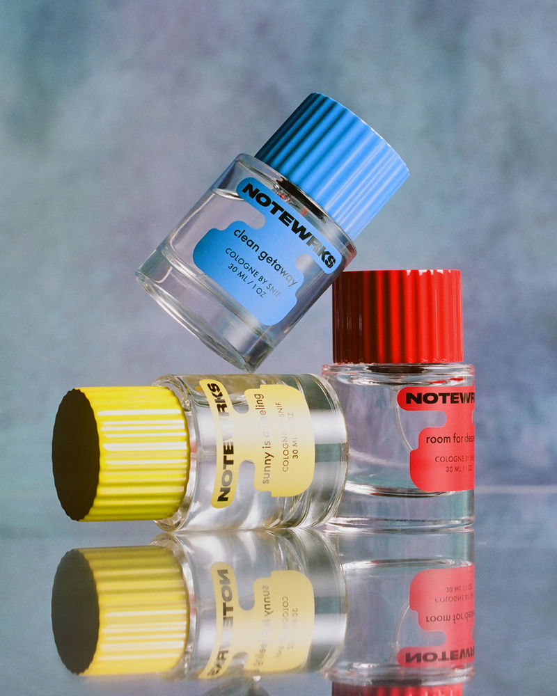 Three NoteWrks cologne bottles with blue ("clean getaway"), yellow ("sunny is calling"), and red ("room for dessert") caps, stacked on a reflective surface.