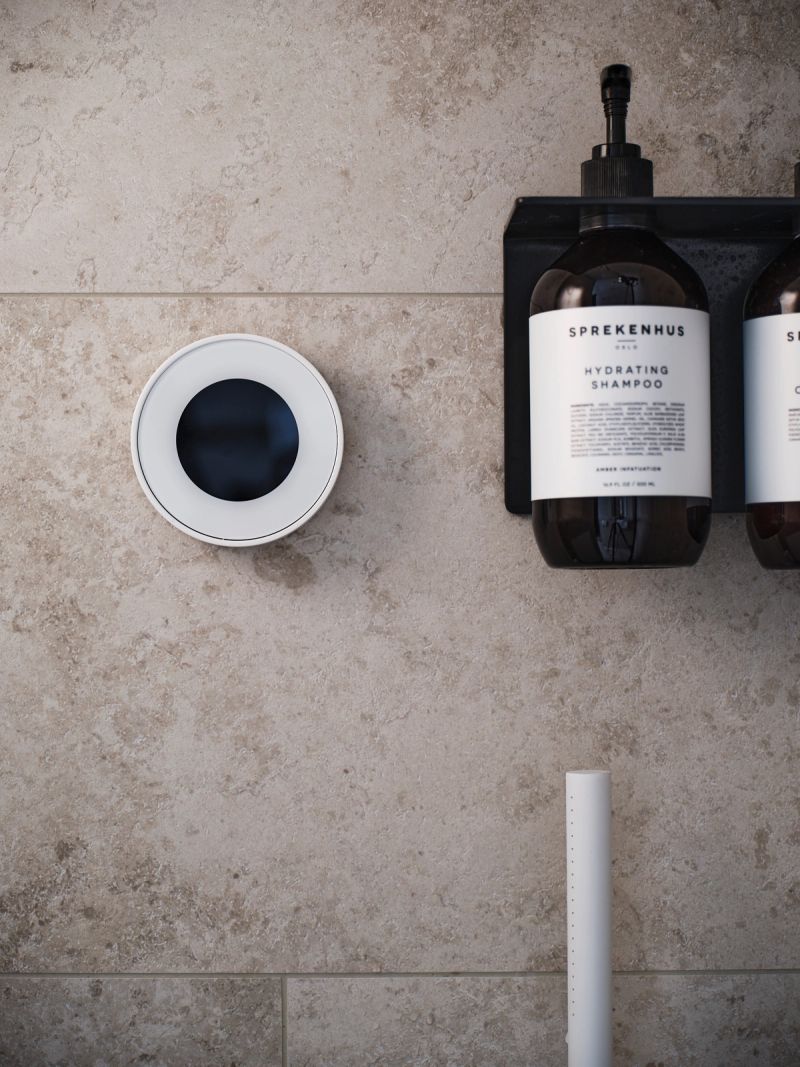 A white circular smart shower controller and dark shampoo bottles on a beige tiled wall.