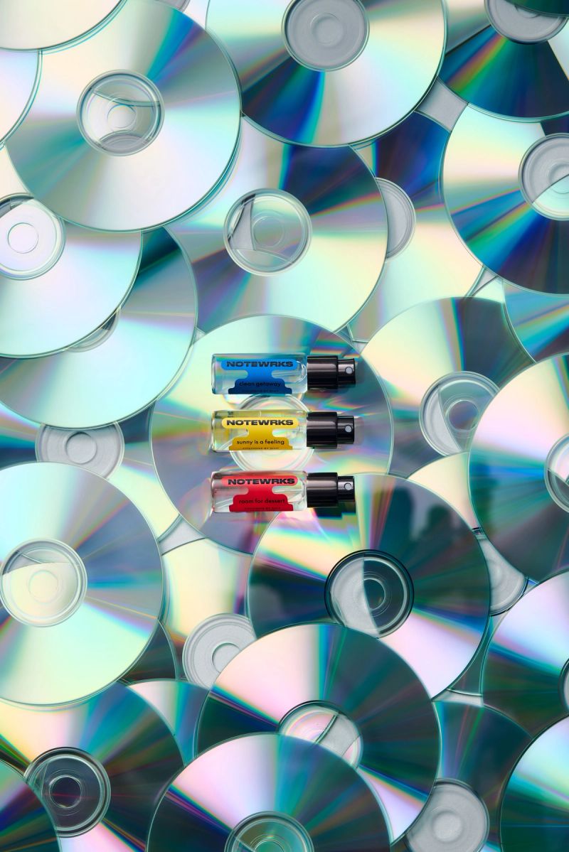 Three NOTEWRKS perfume bottles (blue, yellow, red) on a reflective background of scattered CDs.