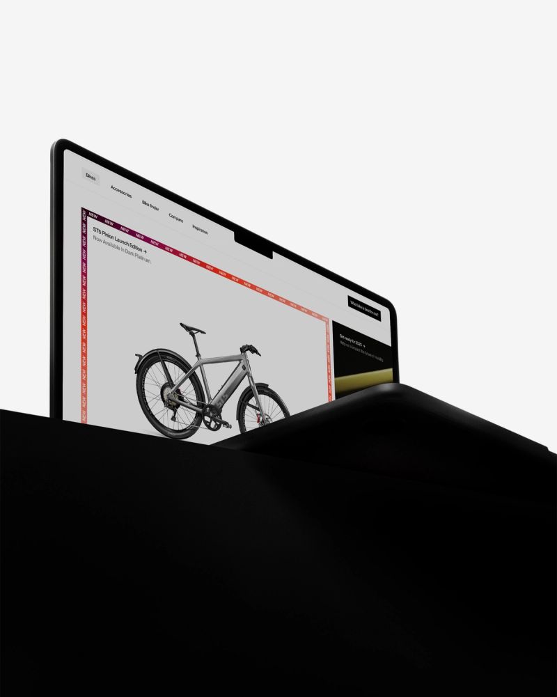Laptop screen displaying a grey electric bike on a website, partially emerging from a dark surface.