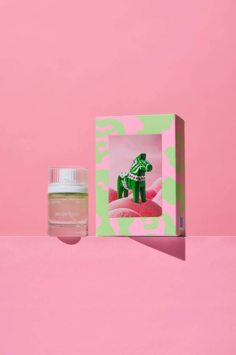 A Swede Tooth perfume bottle next to a pink and green box showcasing a green Dala horse on pink candy, all on a pink background.