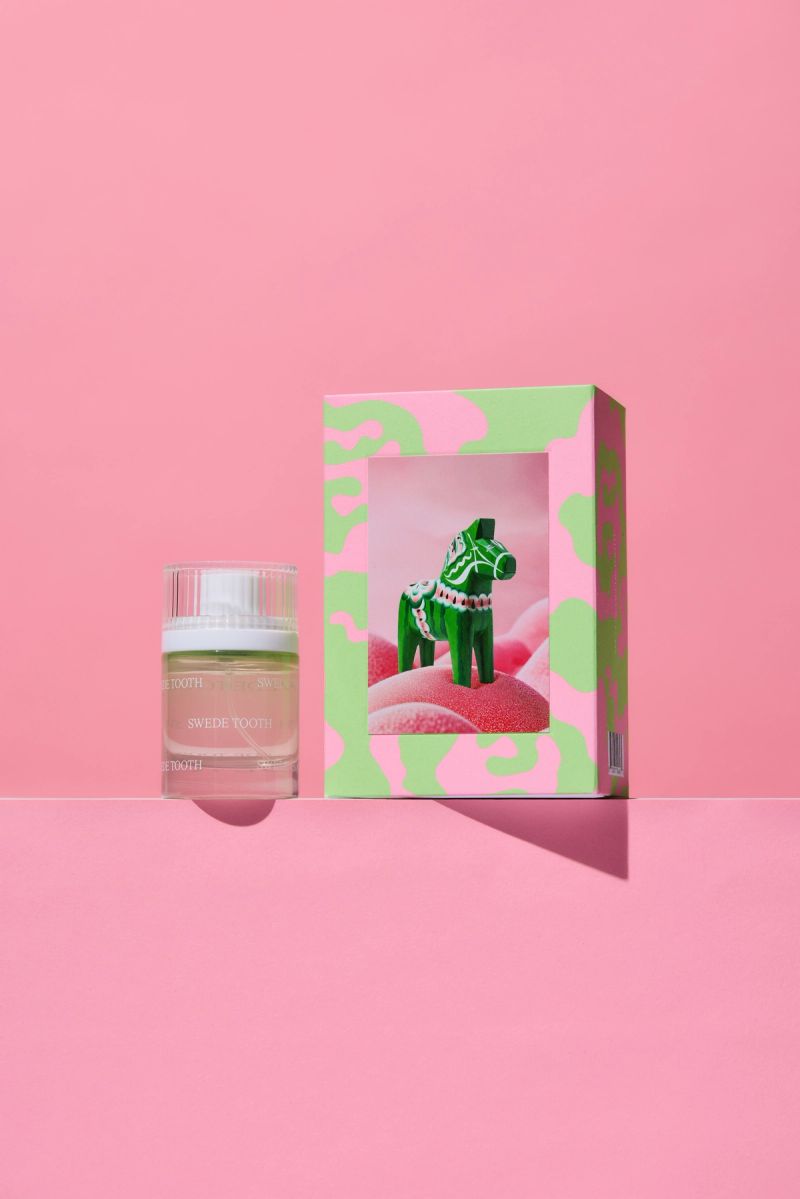 A Swede Tooth perfume bottle next to a pink and green box showcasing a green Dala horse on pink candy, all on a pink background.
