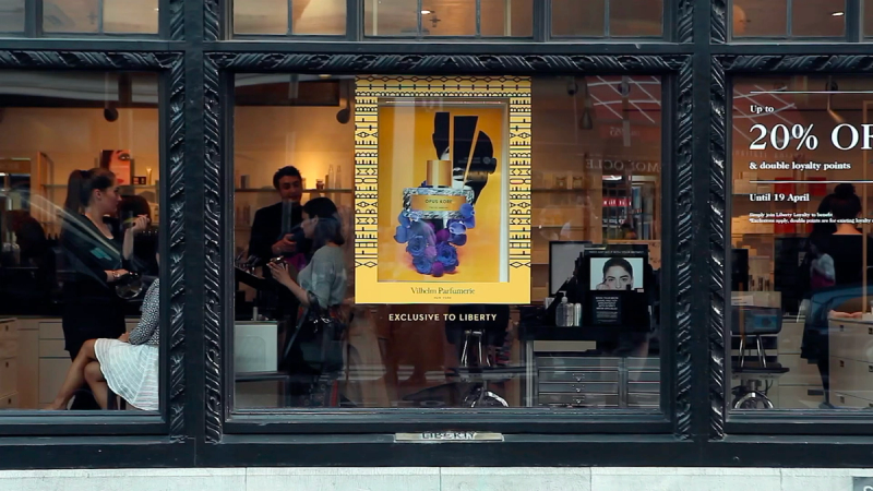 A shop window with reflections, displaying a yellow perfume poster and a sale sign, with people visible inside.