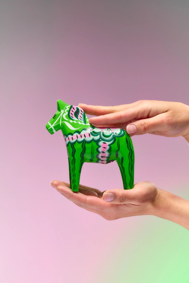 Two hands hold a green Dala horse with white and pink patterns.