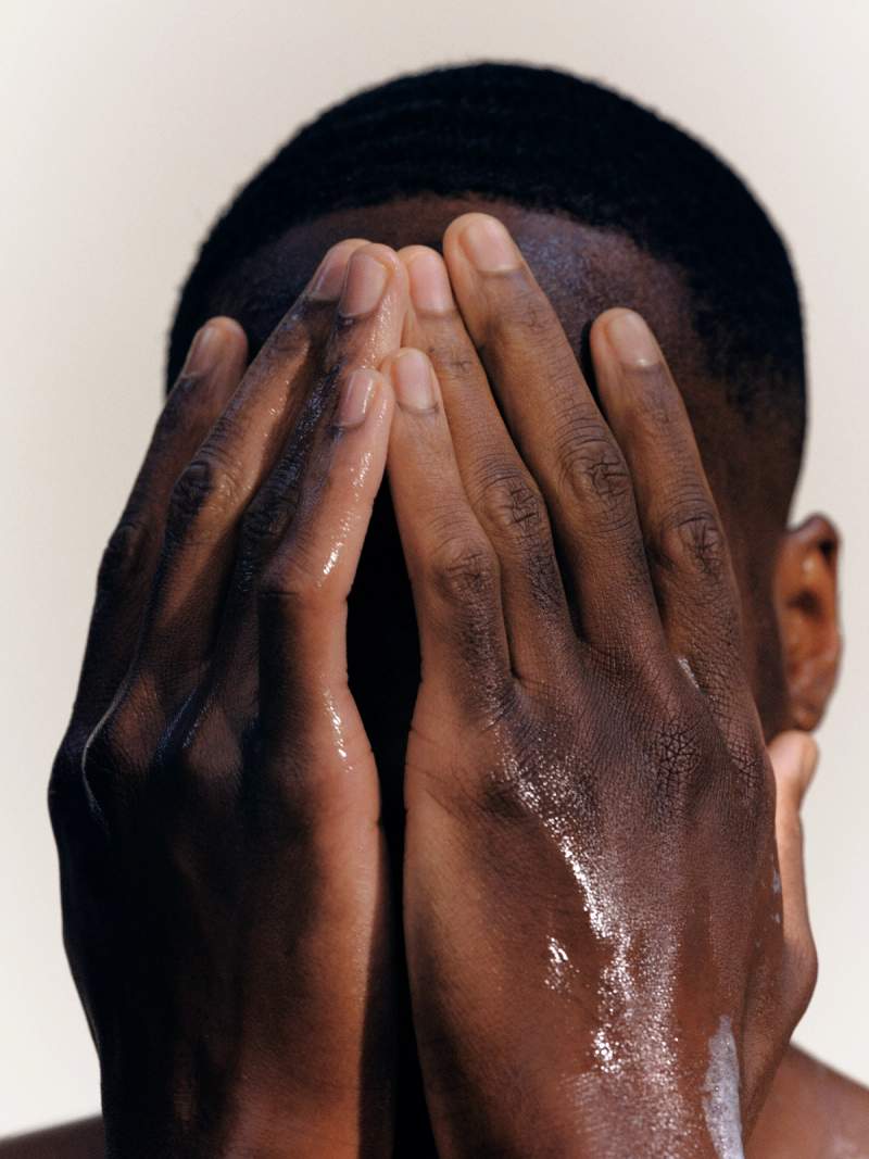A person with dark skin covers their face with wet hands, some white lather visible.