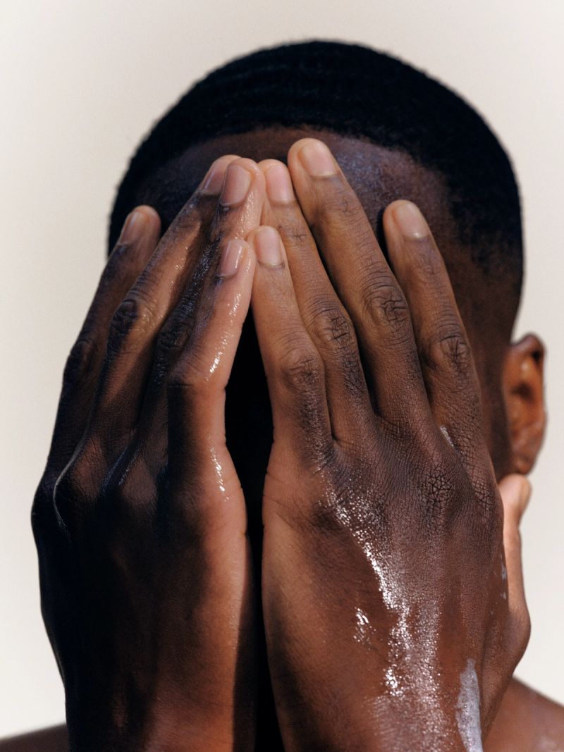 A person with dark skin covers their face with wet hands, some white lather visible.