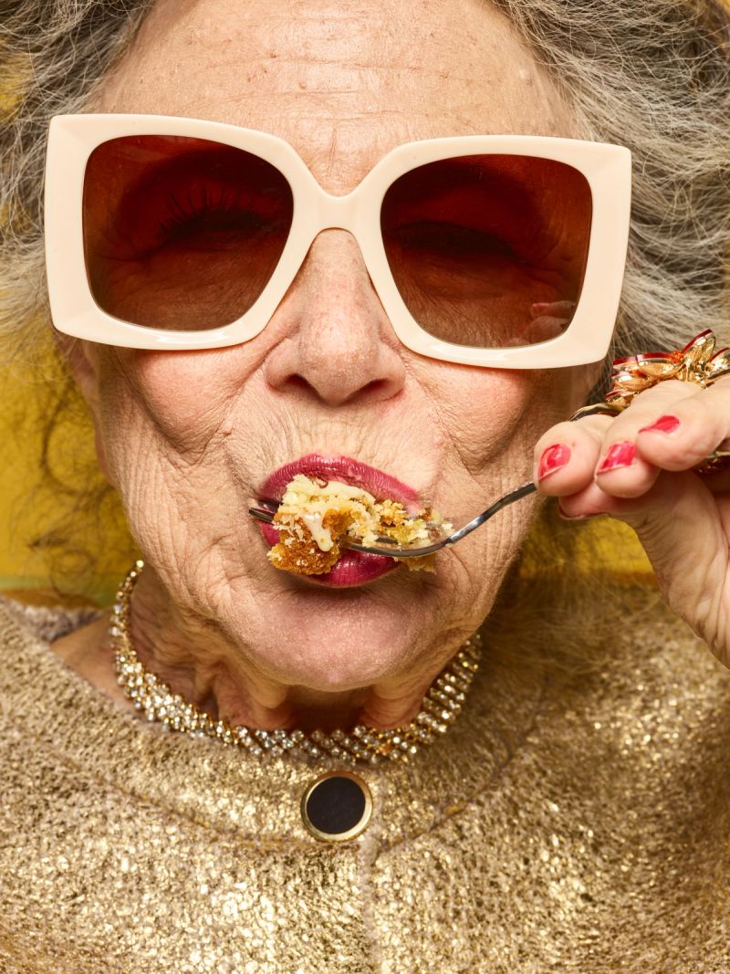 Close-up of a glamorous older woman in oversized sunglasses, a sparkling gold outfit, and red lipstick, eating cake with a fork.