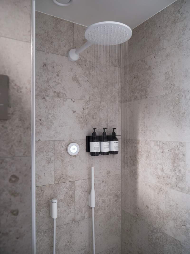 Water streams from a modern rain shower head onto light stone tiles, beside a digital temperature control, wall-mounted toiletries, and two white handheld shower wands.